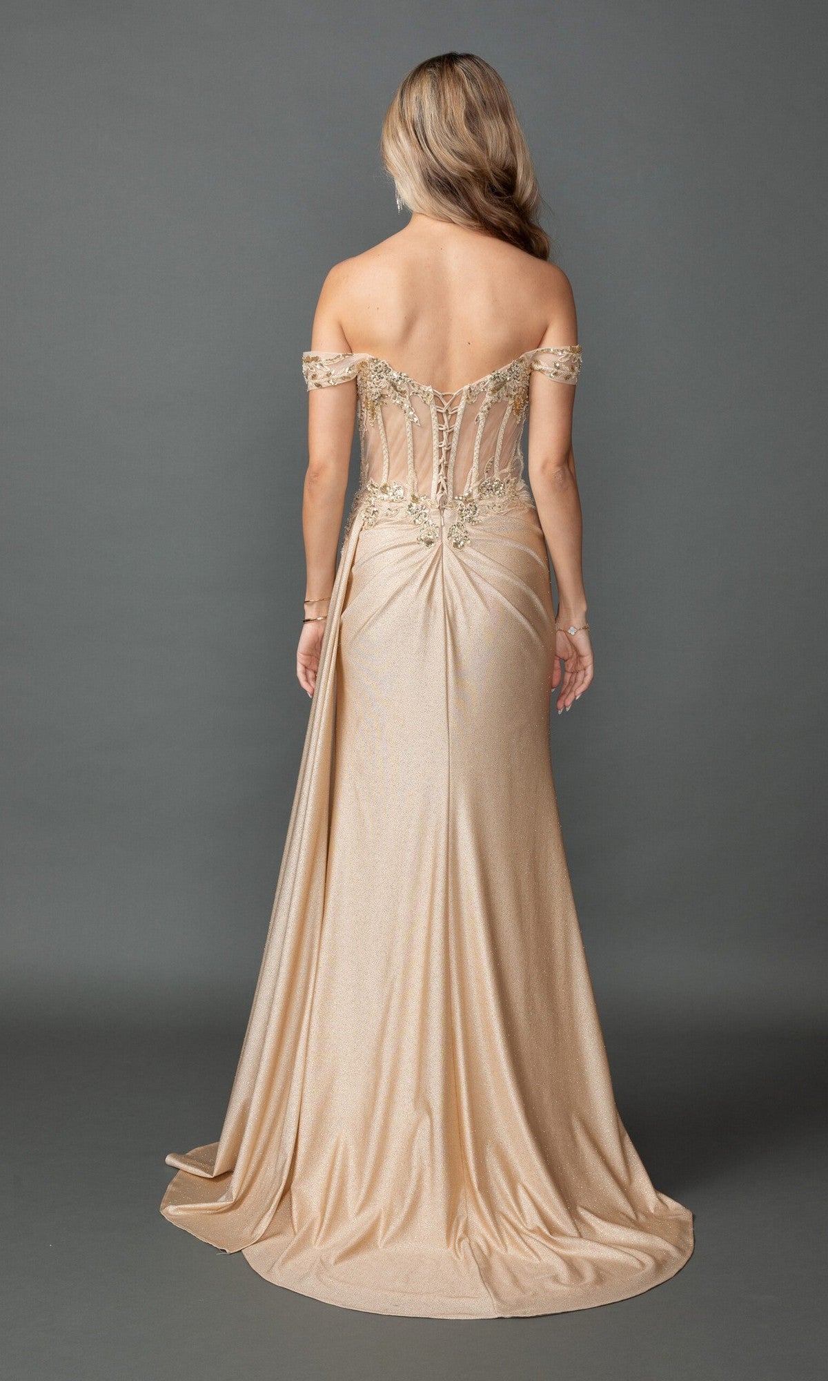 A woman faces away, wearing the Dancing Queen 4535 long prom dress in gold. The off-the-shoulder style features a fitted sheer corset bodice, draped fabric, and elegant lace embroidery. She stands against a plain gray background.